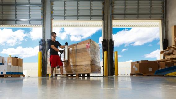 transpalette avec des cartons devant les portes grandes ouvertes d'un espace de chargement de camions. Un homme filme la palette de carton.